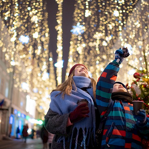 Two people enjoying festive street decorations with warm drinks, surrounded by lights and holiday atmosphere, looking up joyfully.