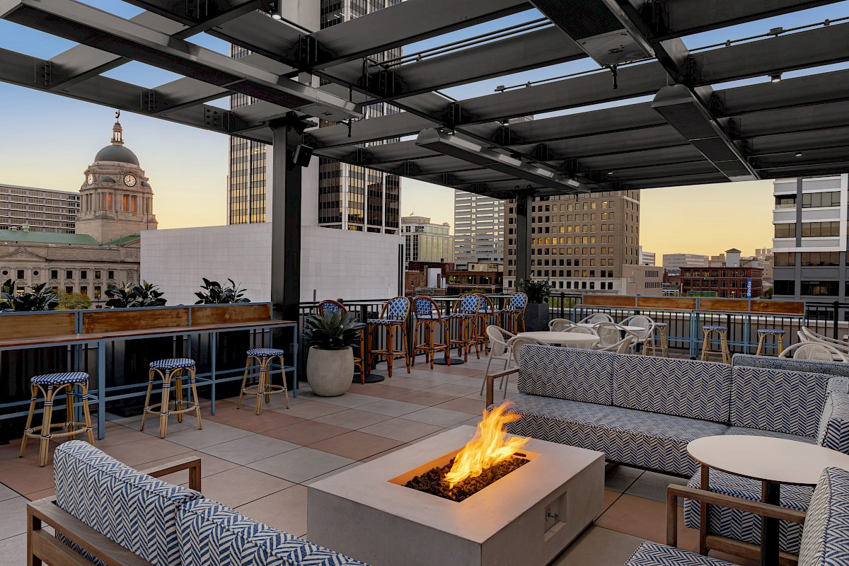 Rooftop lounge with modern seating, a fire pit, and a view of city buildings at sunset. Cozy atmosphere perfect for relaxation and gatherings.