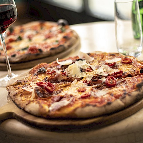 Two pizzas on wooden platters, garnished with cheese and tomatoes, accompanied by a glass of red wine and a green bottle on a table.