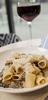 A plate of pasta with sauce, topped with grated cheese sits next to a glass of red wine and a wine bottle on a marble table.