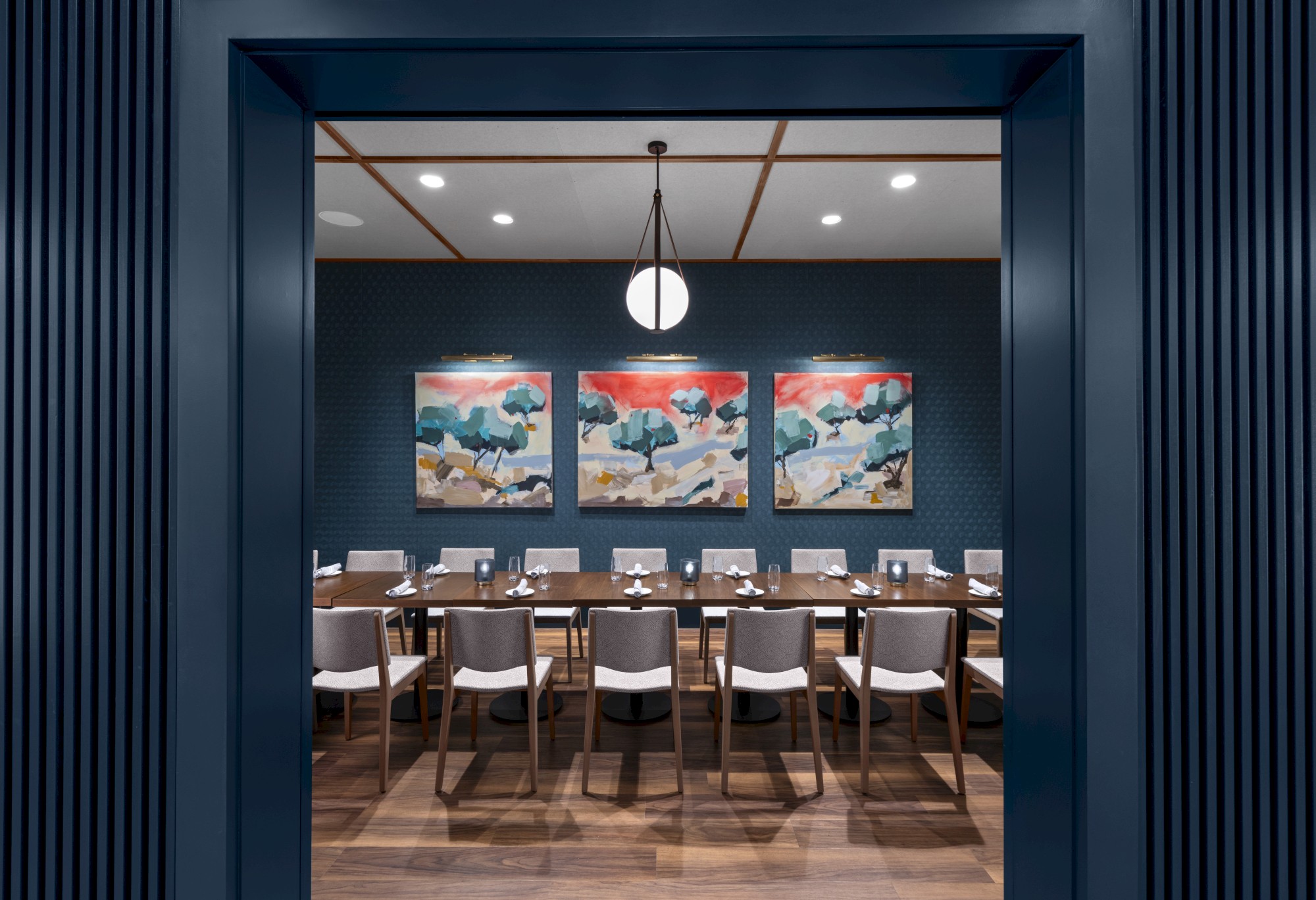 A modern dining room with a long table, chairs, and abstract paintings on the wall, under a central light fixture.