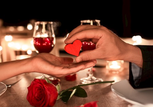 Two hands exchange a red heart over a romantic table setting with roses and wine glasses, creating an intimate atmosphere.