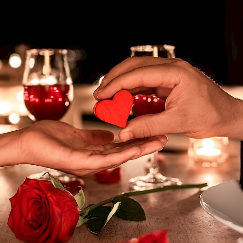 Two hands exchange a red heart over a romantic table setting with roses and wine glasses, creating an intimate atmosphere.