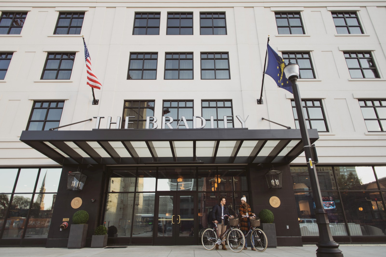 The image shows the entrance of The Bradley, a building with two people standing outside with bicycles, and flags are displayed above.