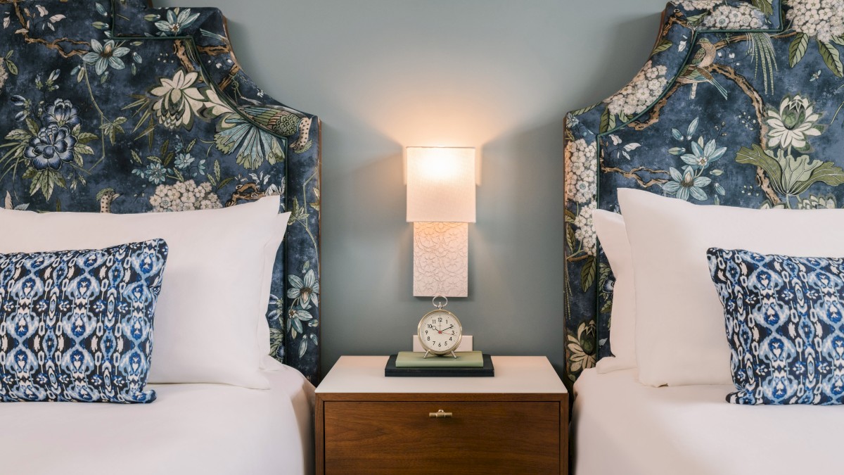 Two beds with floral headboards, a shared nightstand with an alarm clock, and a wall sconce light fixture between them.