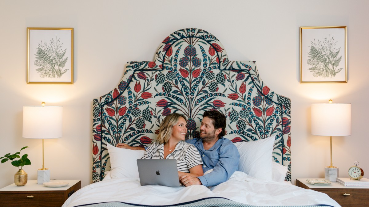 A couple is sitting on a bed with a floral headboard, using a laptop. There are framed plant prints and lamps on bedside tables.