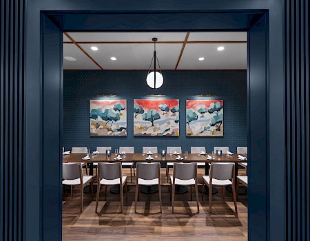 The image shows a dining room with a long table surrounded by chairs, three abstract paintings on the wall, and a round ceiling light.