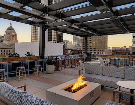 Rooftop lounge with sofas, fire pit, bar stools, and cityscape view. The area is open-air and stylish, perfect for social gatherings.
