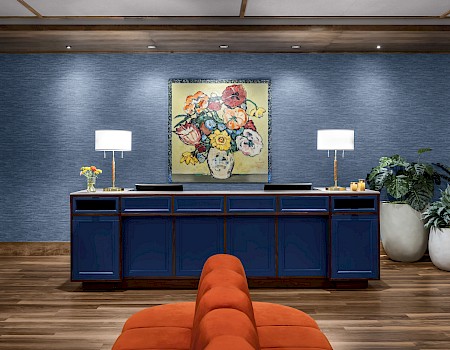 A room with blue walls, a sideboard, table lamps, a colorful painting, potted plants, and an orange cushioned seat on wooden flooring.