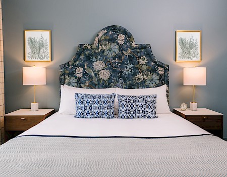A stylish bedroom with a floral headboard, patterned pillows, two lamps on nightstands, and framed botanical art on the wall.