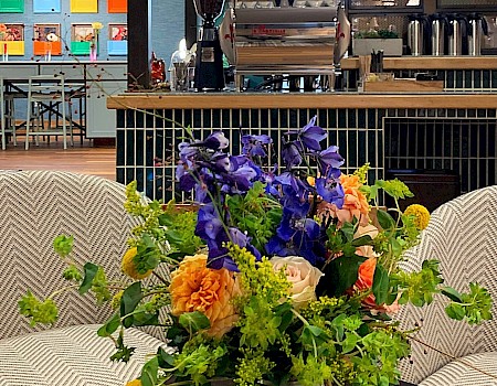 A colorful bouquet sits on a table in a cozy café with an espresso machine and vibrant artwork in the background.