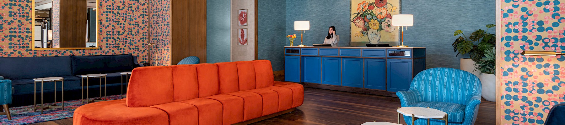 A stylish lounge area with orange and blue seating, floral wallpaper, decorative lighting, and a reception desk in the background.