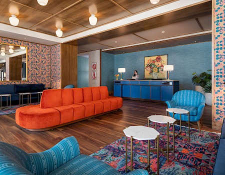 A stylish lounge area with orange and blue seating, floral wallpaper, decorative lighting, and a reception desk in the background.