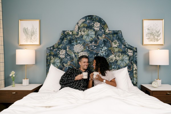 A couple is sitting in bed with coffee, under a floral headboard, and two lamps are on bedside tables with framed art above them.