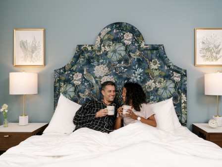 A couple is sitting in bed, smiling and drinking from mugs, with a floral headboard and artwork on the walls.