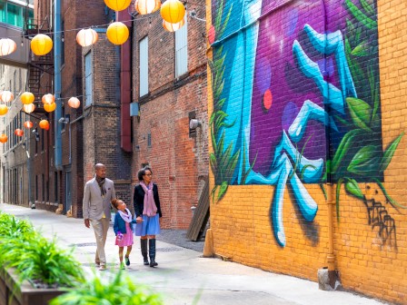 A family walks in an alley adorned with vibrant street art, hanging lanterns, and lush plants along the path.