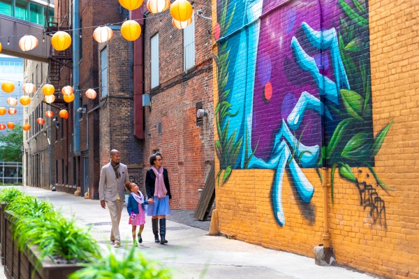 A family walks in an alley adorned with vibrant street art, hanging lanterns, and lush plants along the path.