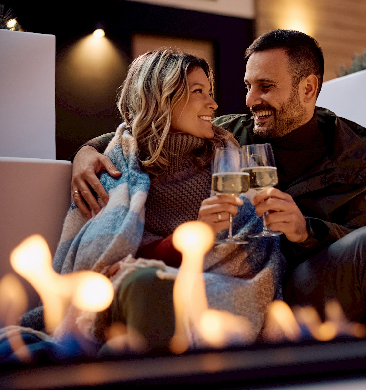 A couple sits closely, wrapped in a blanket, holding wine glasses near a fire pit, enjoying an outdoor setting at night.