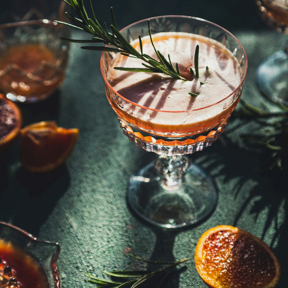 The image shows cocktails in elegant glasses with rosemary garnishes, accompanied by sliced blood oranges on a green surface.