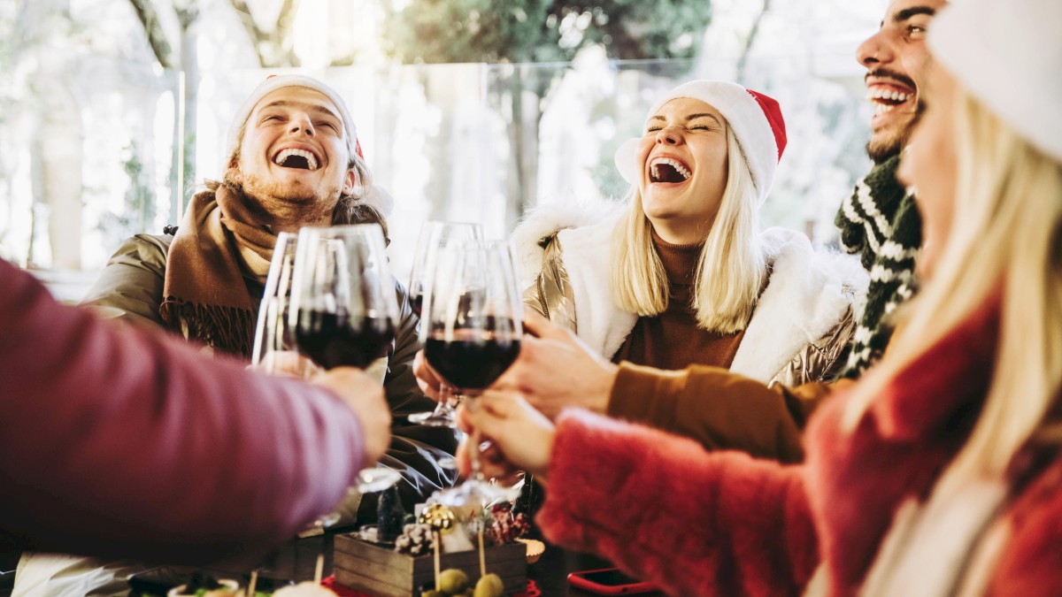 A group of people are joyfully toasting with wine glasses, wearing festive holiday attire and Santa hats, gathered around a table with food.