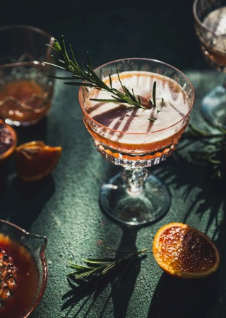 Elegant cocktail with a rosemary garnish in a decorative glass, surrounded by sliced citrus and additional glasses on a green surface.