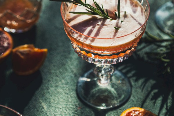 Elegant cocktail with a rosemary garnish in a decorative glass, surrounded by sliced citrus and additional glasses on a green surface.