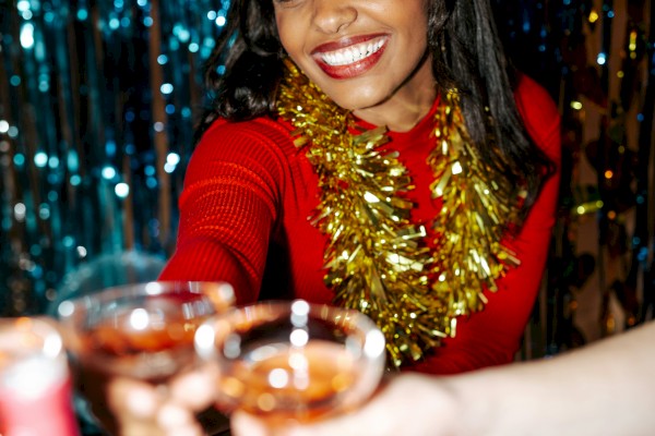 A woman in a red dress, adorned with a gold garland, toasts with others in a festive setting, surrounded by shimmering decorations.