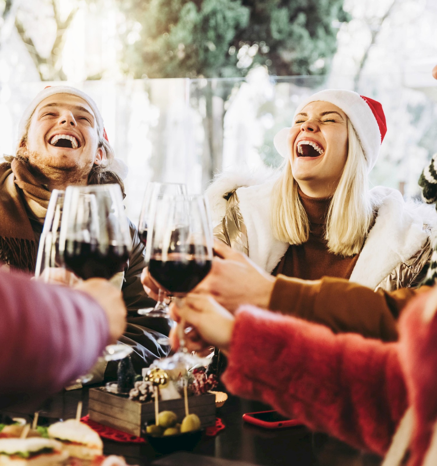 Friends in holiday hats clink wine glasses and laugh around a festive table with snacks, enjoying a cheerful Christmas gathering.