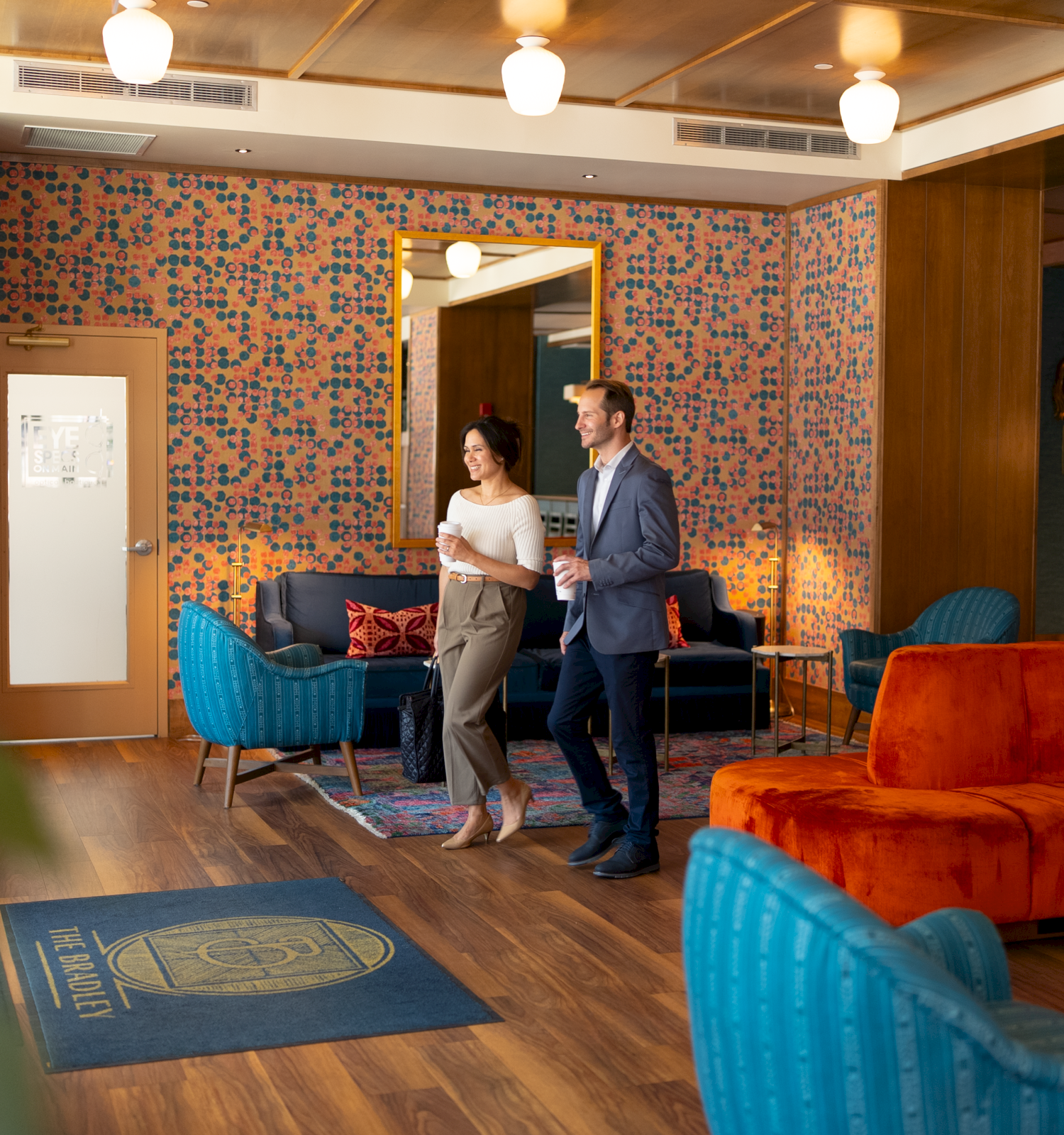 A stylish lobby with blue and orange seating, a patterned wall, two people walking together, warm lighting, and a blue rug at the entrance.