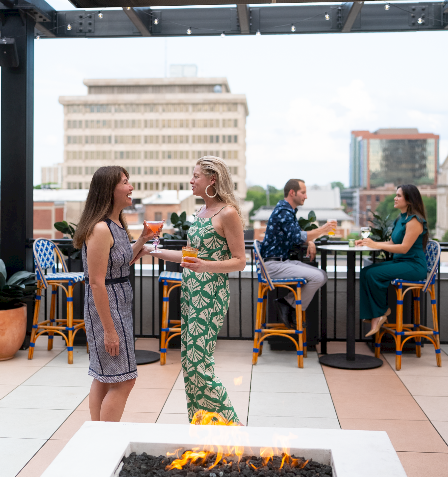 Two women chat by a rooftop fire pit with cocktails, while two others sit at a high table in the background; urban skyline behind them.