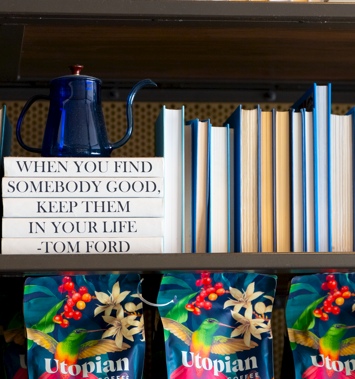 A shelf with books, a blue kettle, and colorful Utopian-themed bags; a Tom Ford quote sign reads: “When you find somebody good, keep them in your life.”