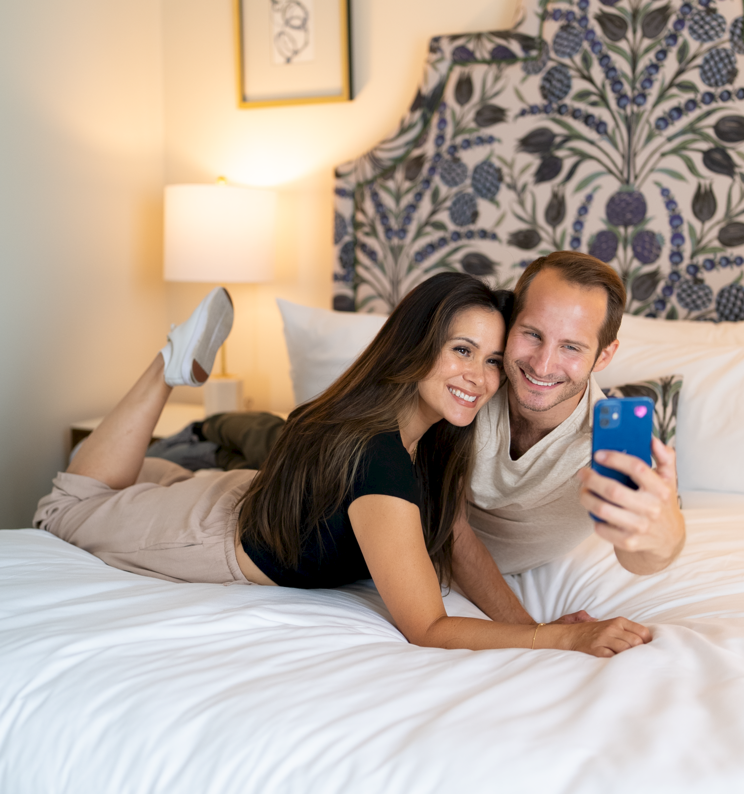 Two smiling people lie on a white bed, taking a selfie with a blue phone, in a cozy bedroom with a patterned headboard and soft lighting.