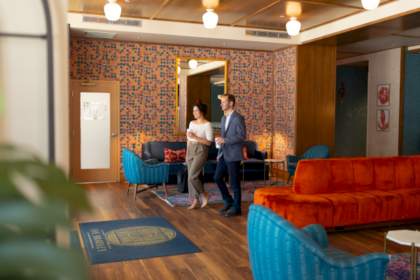 Two people walk through a modern, colorful lounge with blue and orange seating, warm wood floors, and patterned wall tiles.
