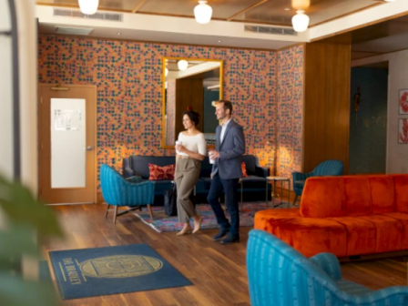 Two people walk through a stylish, cozy lounge with colorful chairs, a wooden floor, and a bright orange sofa, as a patterned wall and plants frame the scene.