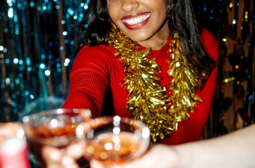A woman in a red dress with gold garland smiles at a party, toasting glasses of champagne against a shiny backdrop.