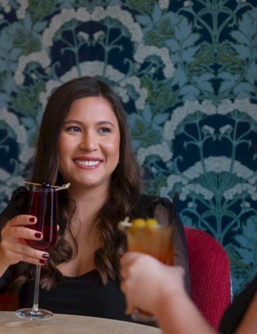 Two women enjoy drinks at a table, smiling and toasting with wine and a cocktail against a patterned blue-green wall.