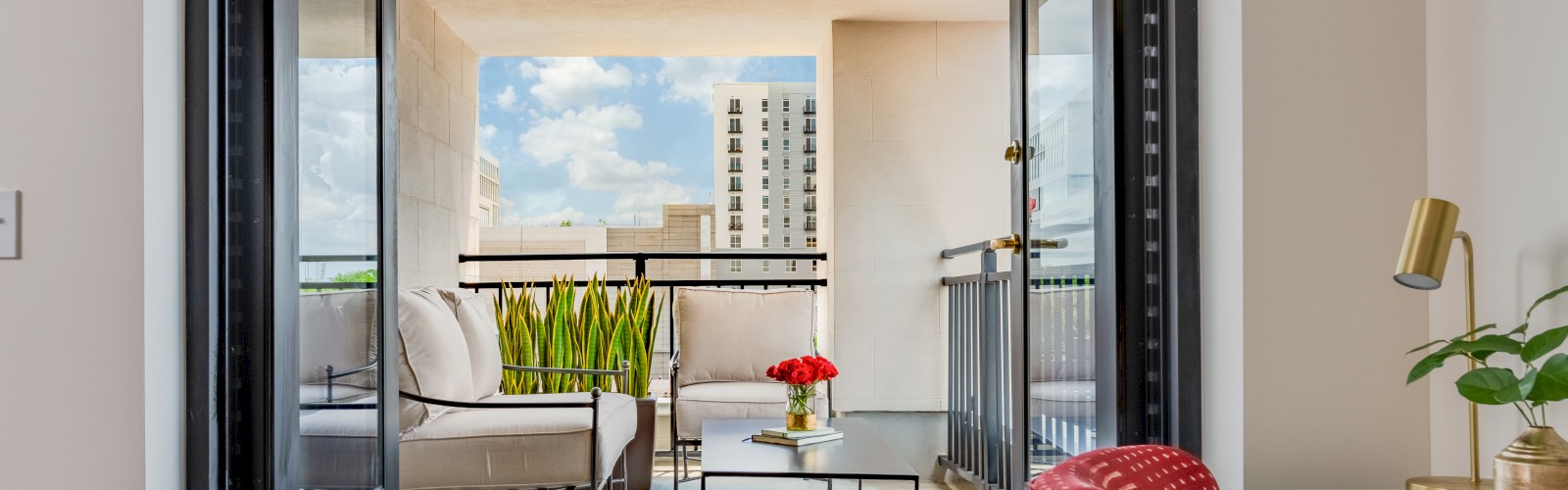 This is a bright, modern balcony with outdoor seating, potted plants, and a city skyline view under a blue sky.