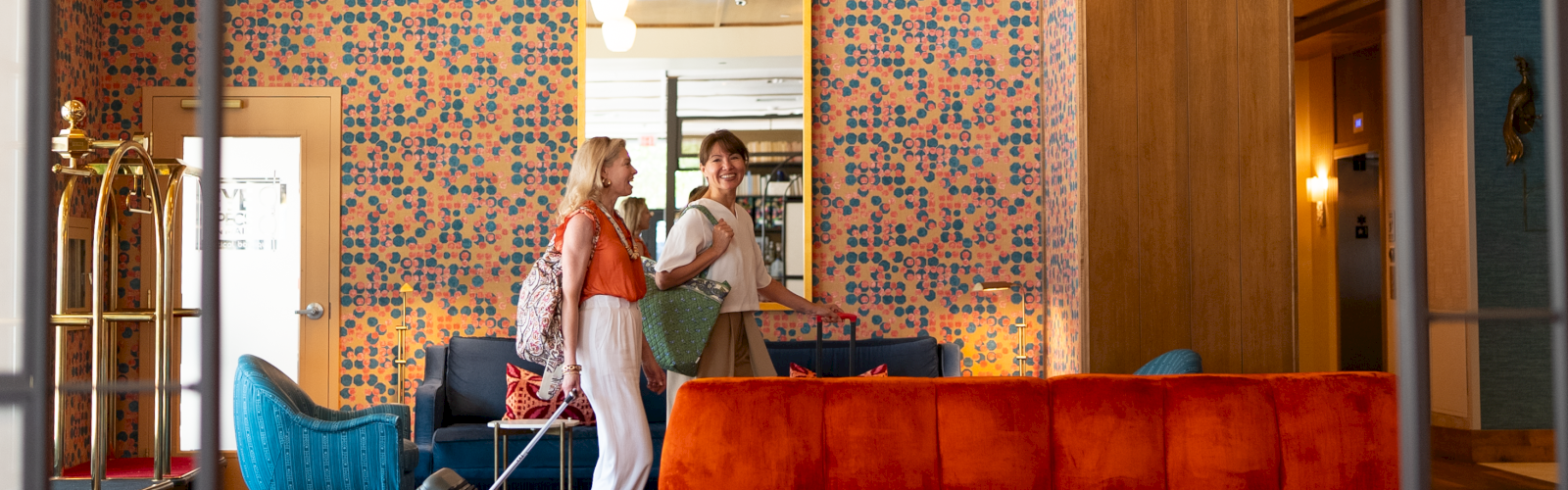 Two women with luggage are talking and smiling in a colorful, stylish hotel lobby with orange, blue, and patterned decor.
