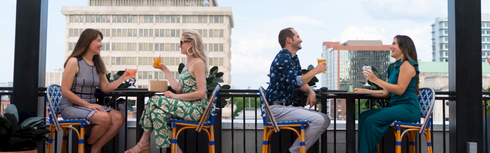 Four women and two men sit on bar stools, drinking cocktails and chatting on a rooftop with city buildings in the background.