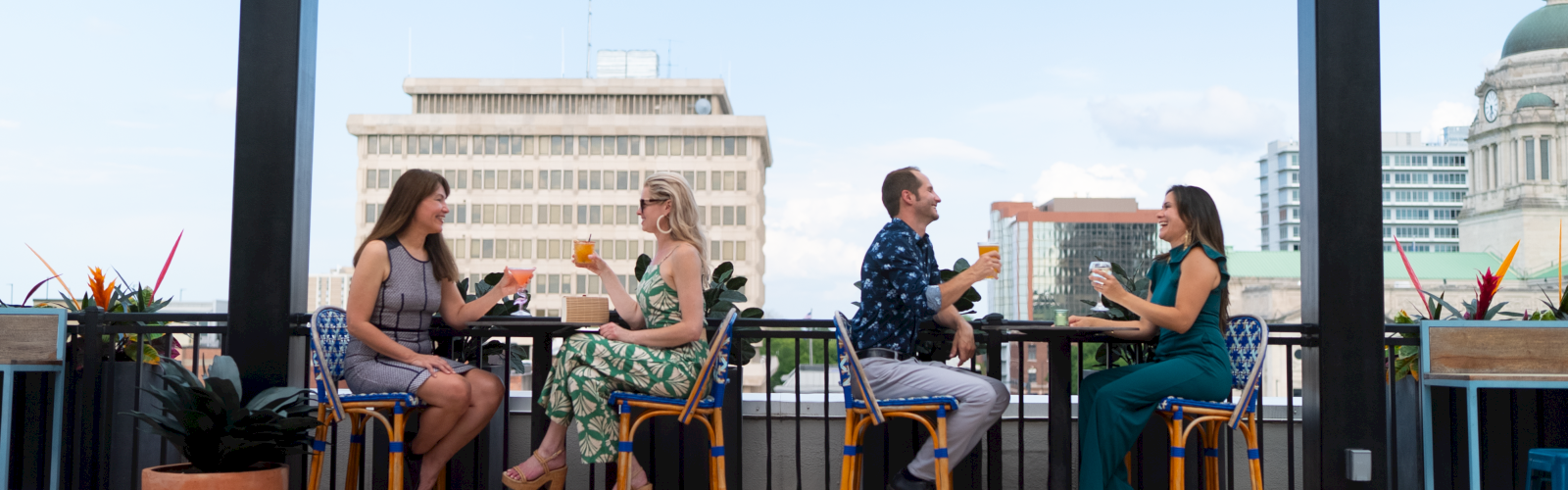 Four couples are enjoying drinks and conversation on a rooftop patio with city buildings in the background.