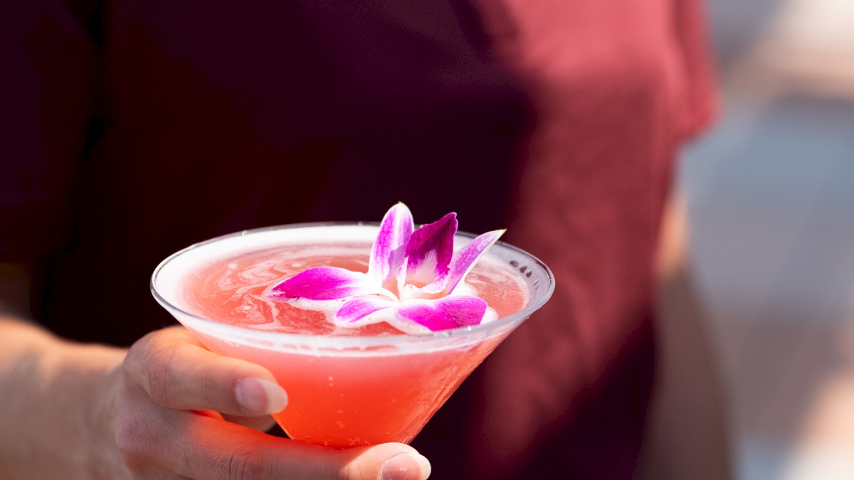 A person is holding a pink cocktail garnished with pink and white flowers.