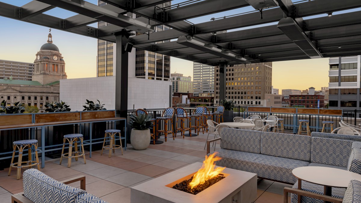 The image shows a rooftop lounge with seating, plants, and a fire pit, set against a city skyline at sunset.