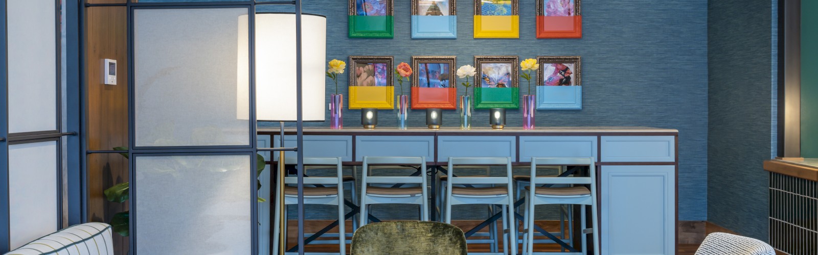 A cozy lounge area with a patterned sofa, colorful chairs, framed artwork, and a bar with vibrant decorations in the background.